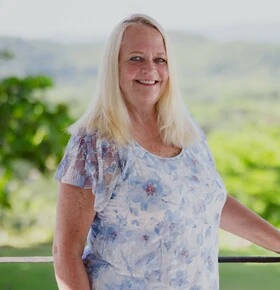 Woman with long light hair in a blue floral blouse standing on a terrace with green hills behind